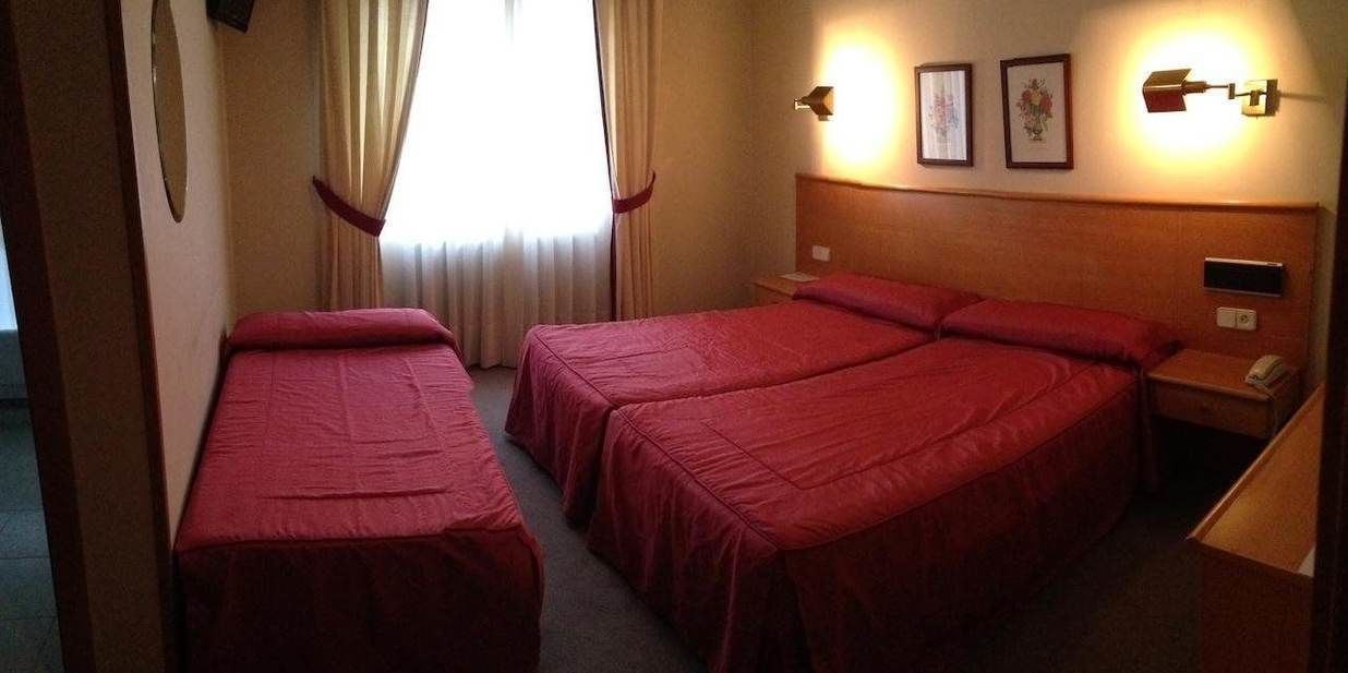 Ein Hotelzimmer mit zwei Einzelbetten und einem größeren Bett. Ein Nachttisch mit Lampe und Telefon ist vorhanden. Das Zimmer verfügt über ein Fenster mit Vorhängen und einer Wandleuchte.