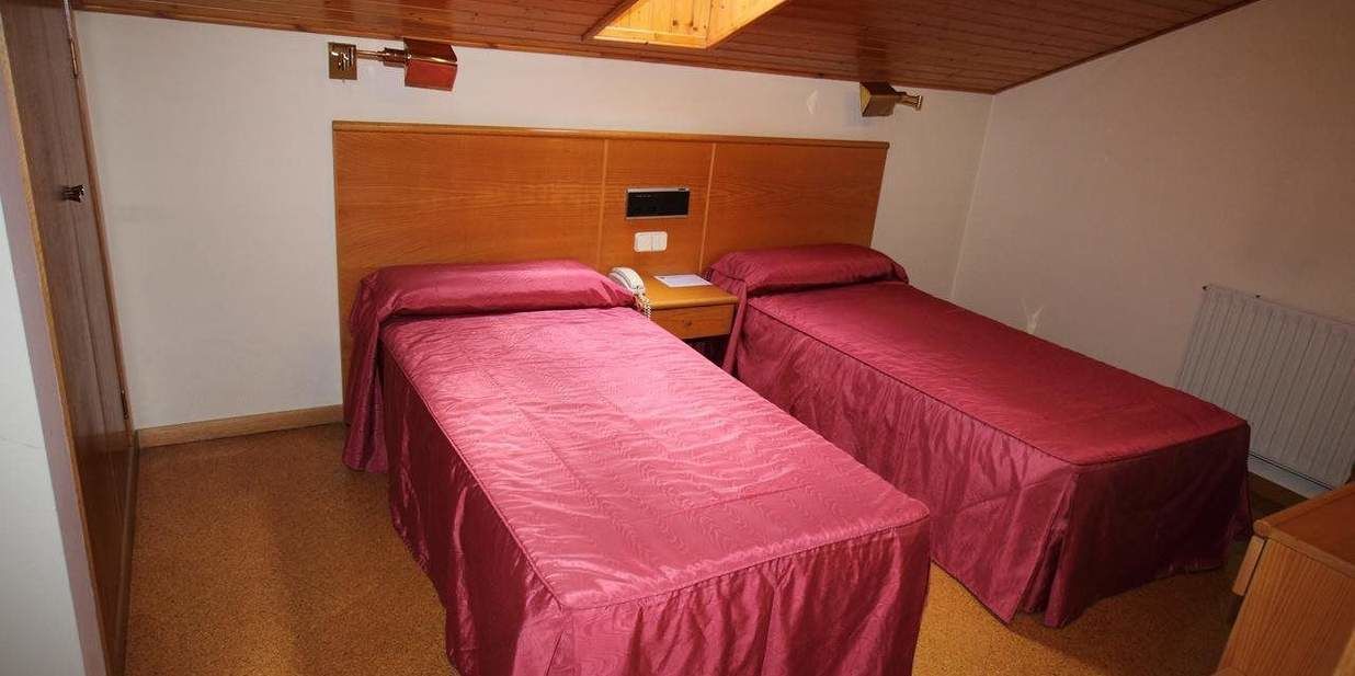 Een kleine kamer met twee bedden voorzien van rode beddengoed, een houten hoofdbord, een klein tafeltje met een telefoon en een radiator aan de muur.