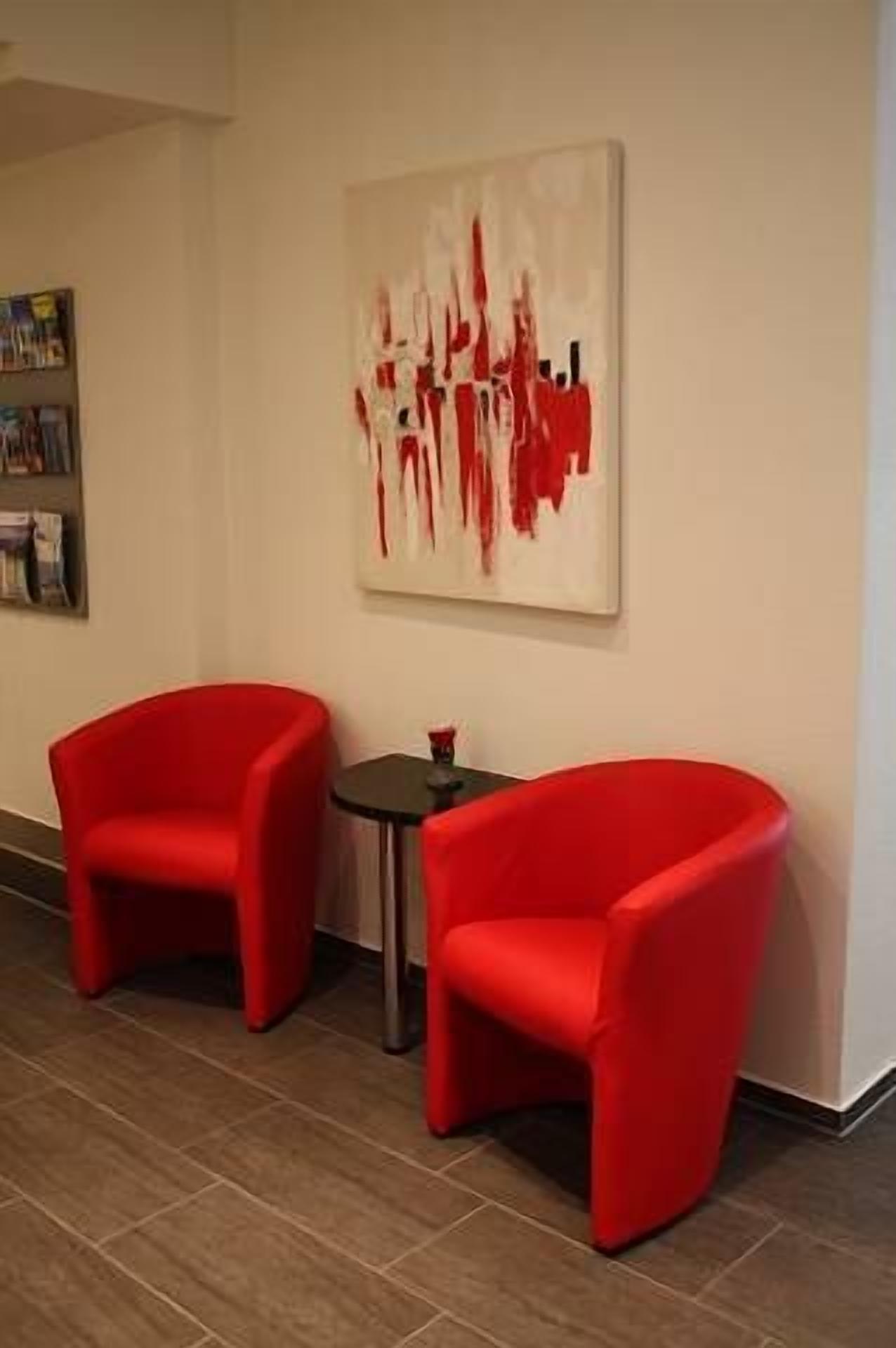 Une pièce moderne avec deux fauteuils rouges et une petite table, une grande peinture abstraite au mur et une bibliothèque remplie de livres.