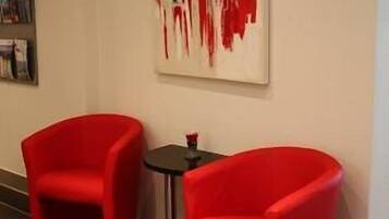 Une pièce moderne avec deux fauteuils rouges et une petite table, une grande peinture abstraite au mur et une bibliothèque remplie de livres.
