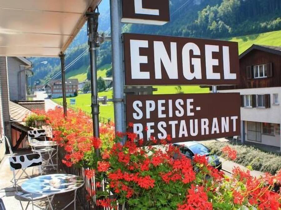 En restaurangskylt med namnet ”ENGEL SPEISE-RESTAURANT” och en vy över en stad med hus och grönska i bakgrunden.