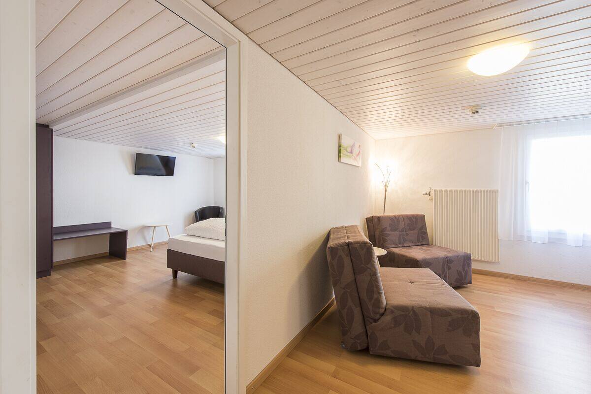 A modern, well-lit room with a bed, two armchairs, a small table, and a TV mounted on the wall.