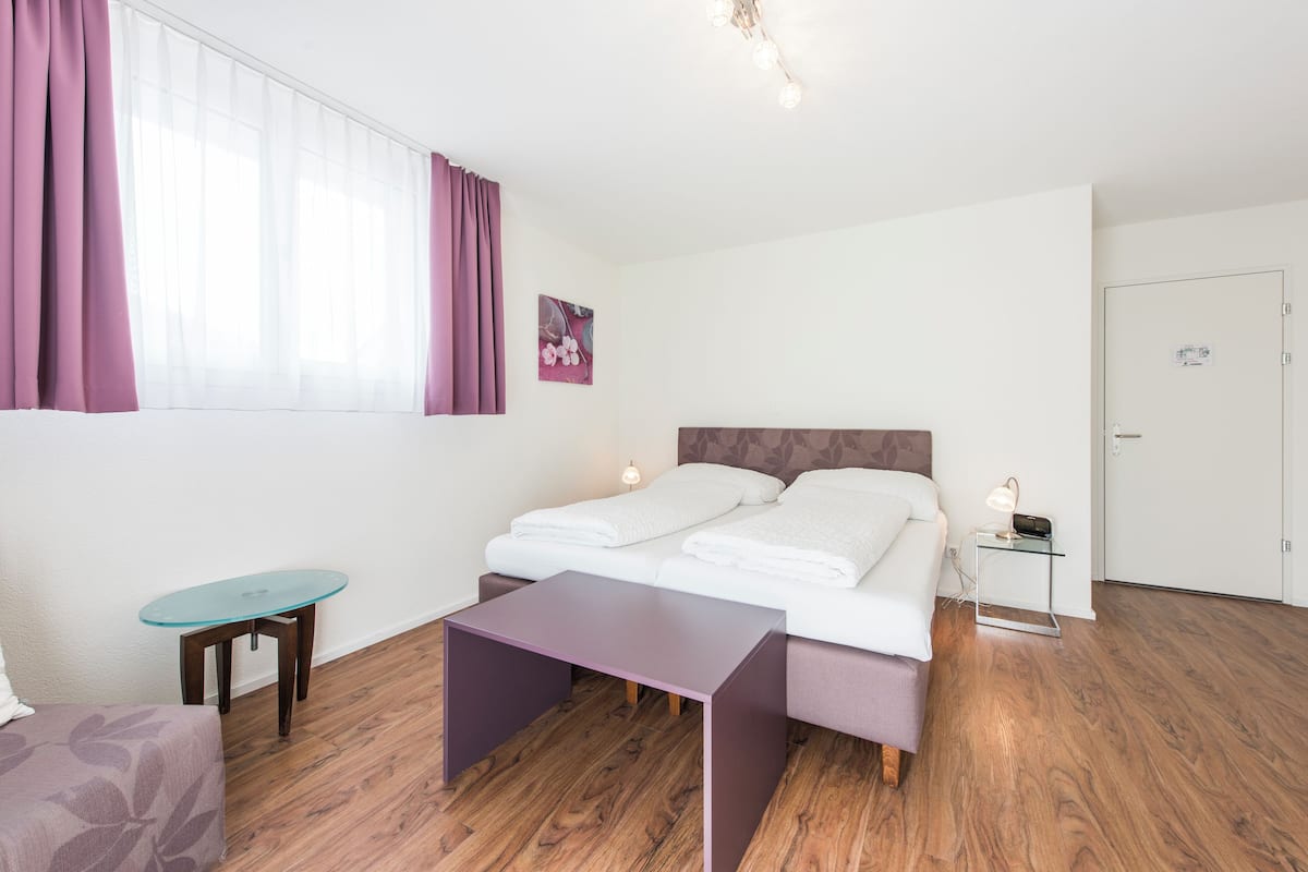 A bedroom with a bed, a small table, a nightstand, a window with curtains, and a door.