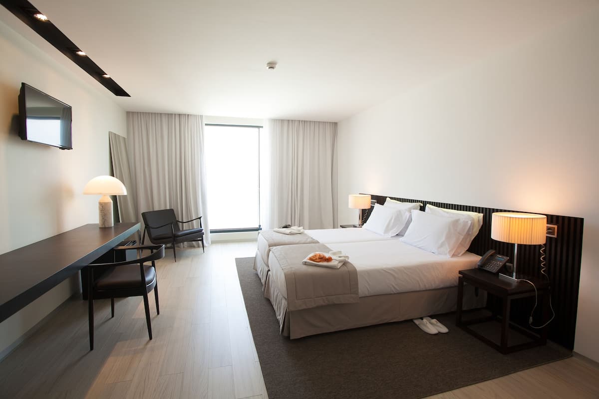 A hotel room with a bed, bedside tables, a desk, a chair, and a television on the wall.