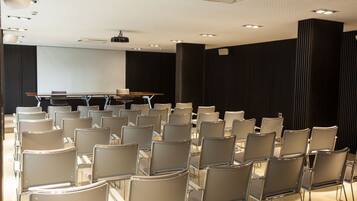 Un salón de conferencias con filas de sillas, una pantalla de proyector y una mesa.