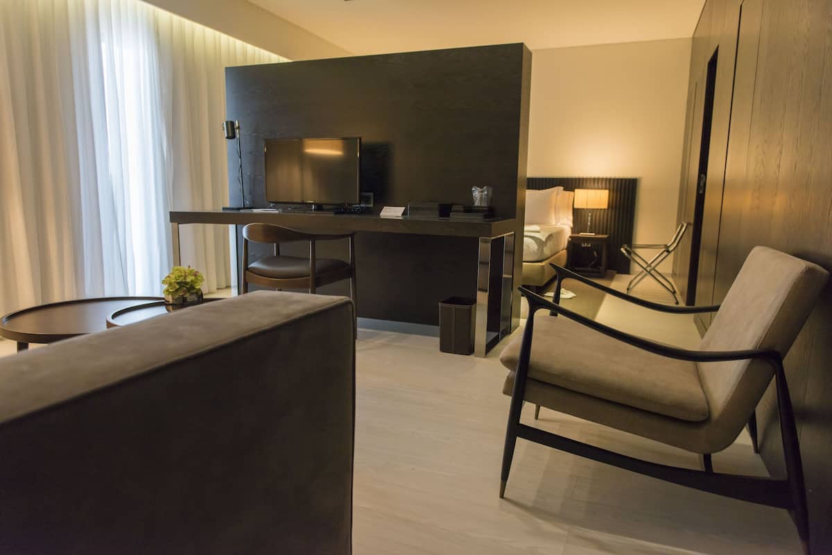 A modern hotel room with a sofa, armchair, small round table, and a desk with a chair.