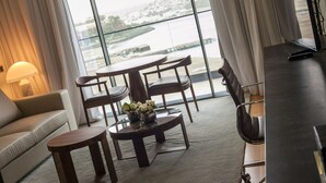 Habitación de hotel moderna con un ventanal grande que ofrece vistas a un río y al paisaje urbano. Hay un sofá, una mesita redonda con un florero y una mesa de comedor con sillas.