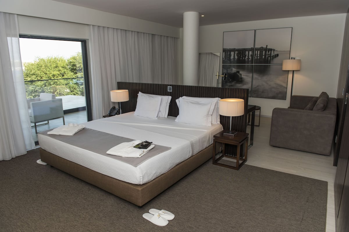 A modern hotel room with a large bed, a sofa, a small table, and a view of greenery outside.