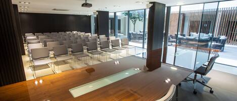 A modern conference room with a wooden table, rows of chairs, and large windows offering a view of outdoor seating.