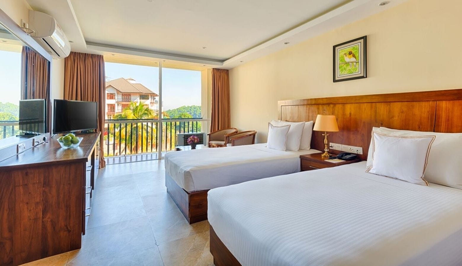 A hotel room with two beds, a desk with a TV, a balcony with a view of a building and trees.