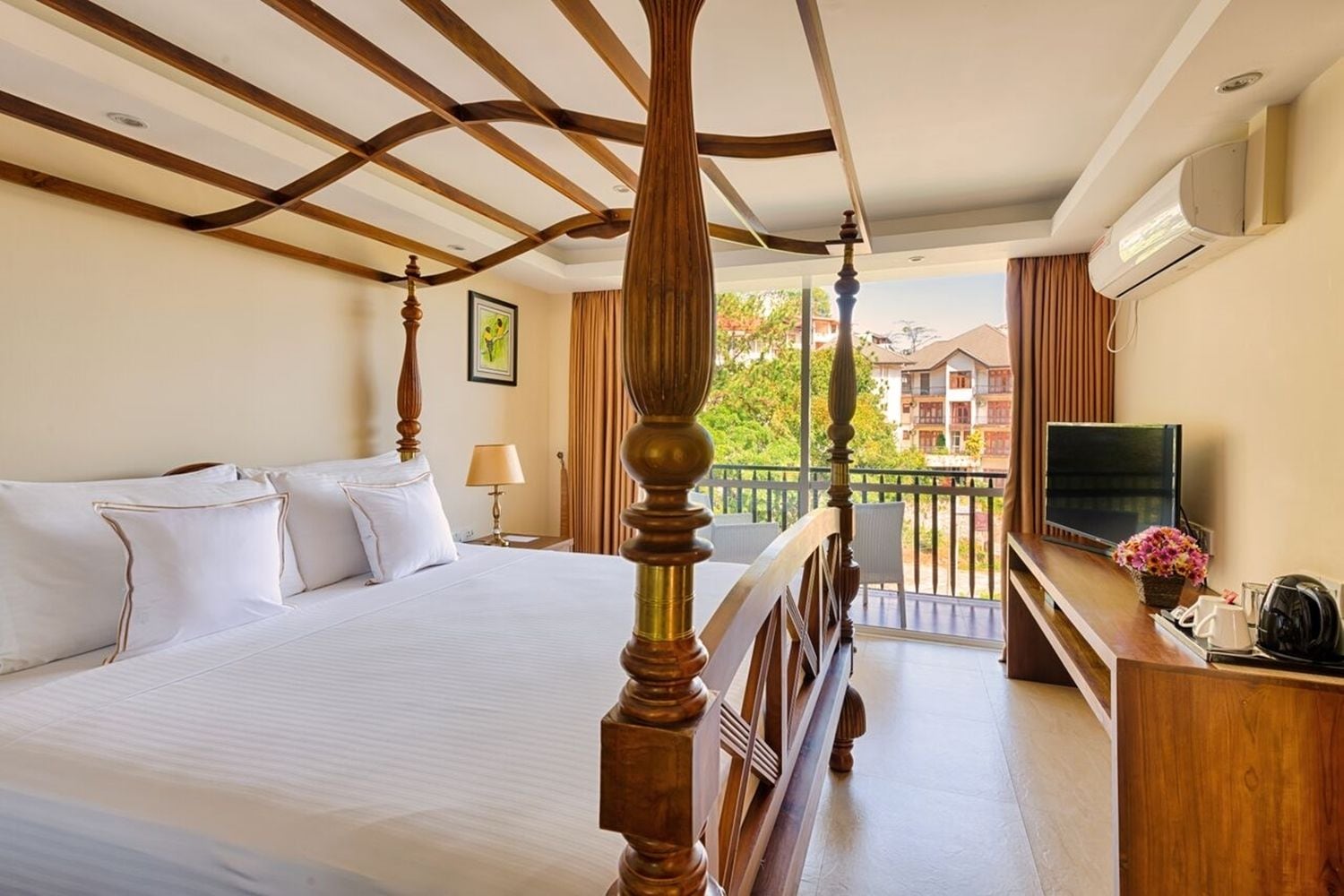A hotel room with a four-poster bed, a television, a desk with a coffee maker, and a balcony with a view of buildings.