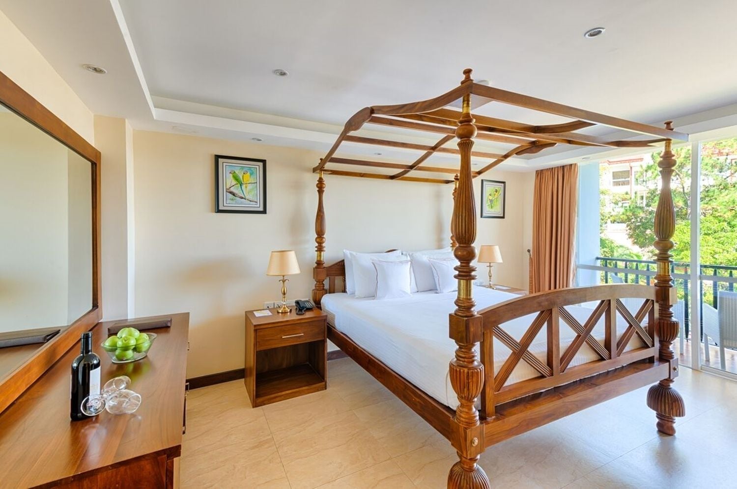 A four-poster bed in a hotel room with a wooden headboard, a nightstand with a lamp, a small table with a vase and apples, and a view of the outdoors through large windows.