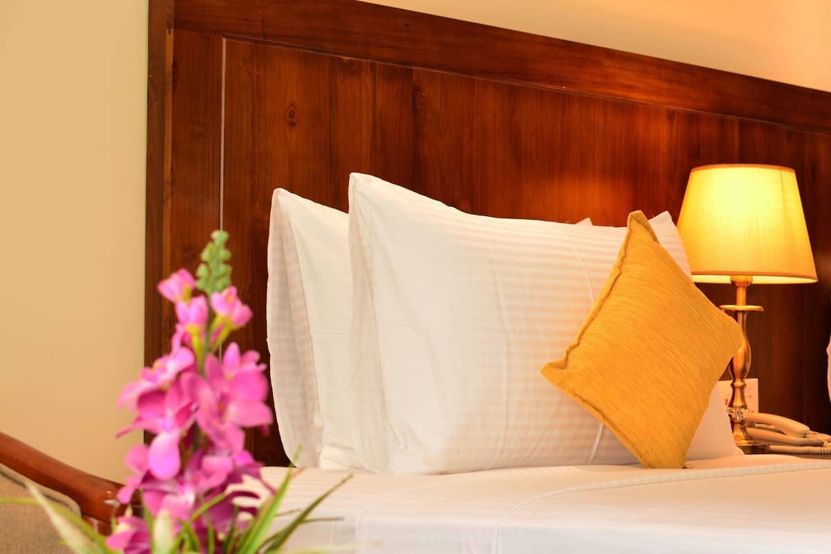 Quarto de hotel com cabeceira de madeira, roupa de cama branca, uma almofada amarela, abajur e planta em vaso com flores rosa.