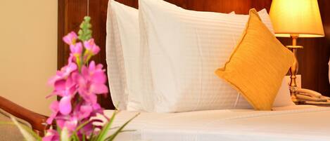 Quarto de hotel com cabeceira de madeira, roupa de cama branca, uma almofada amarela, abajur e planta em vaso com flores rosa.