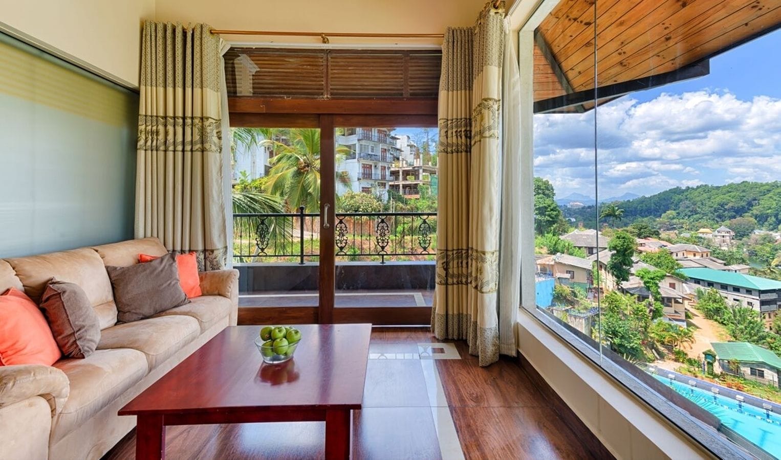 A room with a sofa, a wooden table with a bowl of fruit, and a large window with a view of a residential area and a pool.