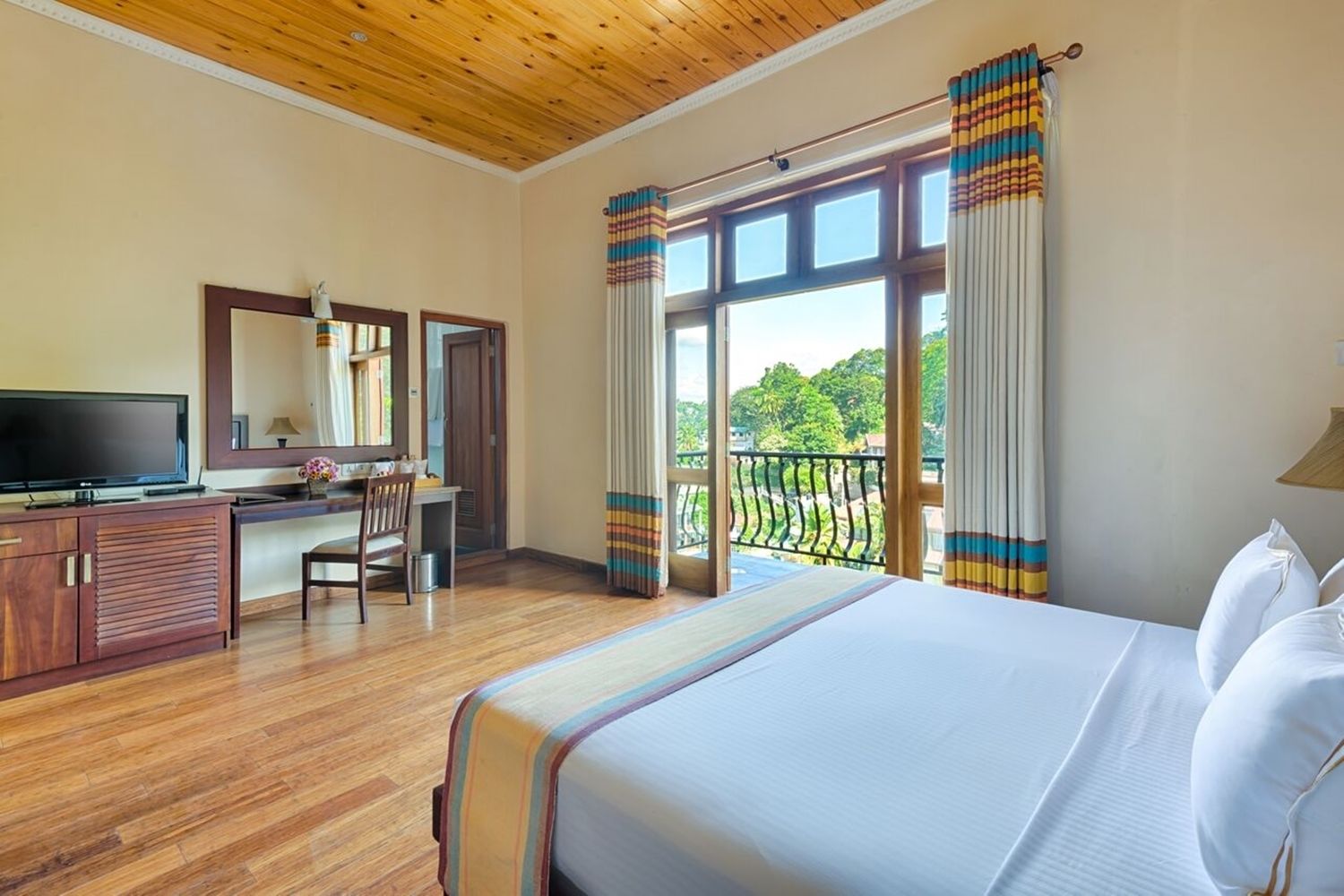 Habitación de hotel con techo de madera, cama con ropa blanca, escritorio con televisor y balcón con cortinas.