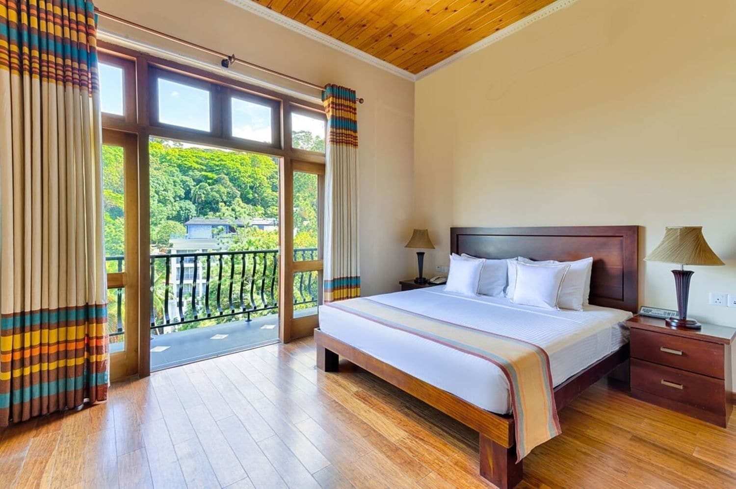 Une chambre d’hôtel avec un grand lit, un plancher en bois, une table de chevet avec une lampe et une vue sur la verdure, visible à travers les portes du balcon.