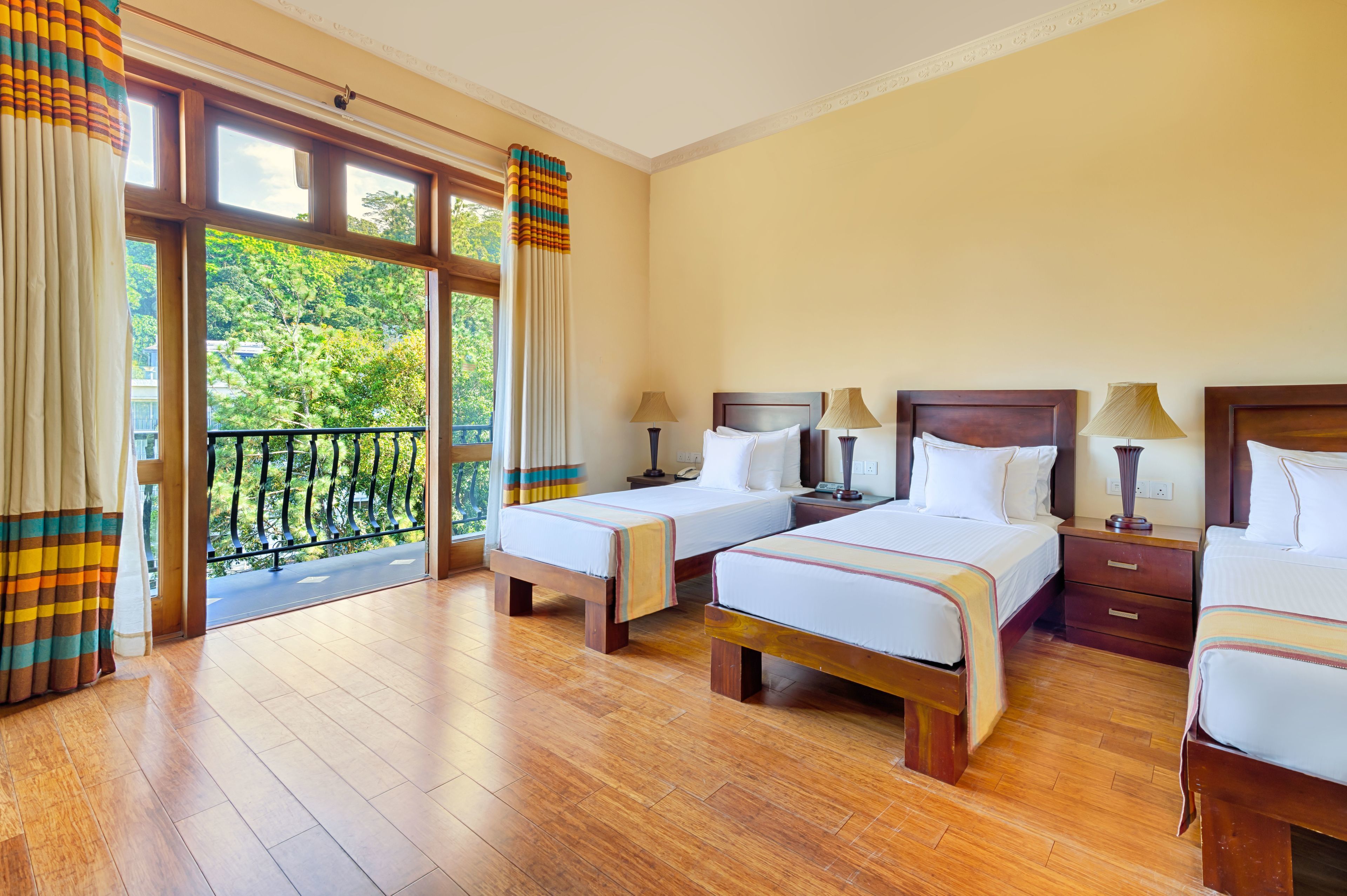 A hotel room with two beds, wooden flooring, and a balcony with a view of trees.