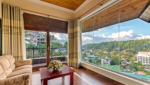 A spacious room with a large window offering a view of a residential area and hills. A sofa and a wooden coffee table with a floral arrangement are visible. - Serene Kandy (Kandy)