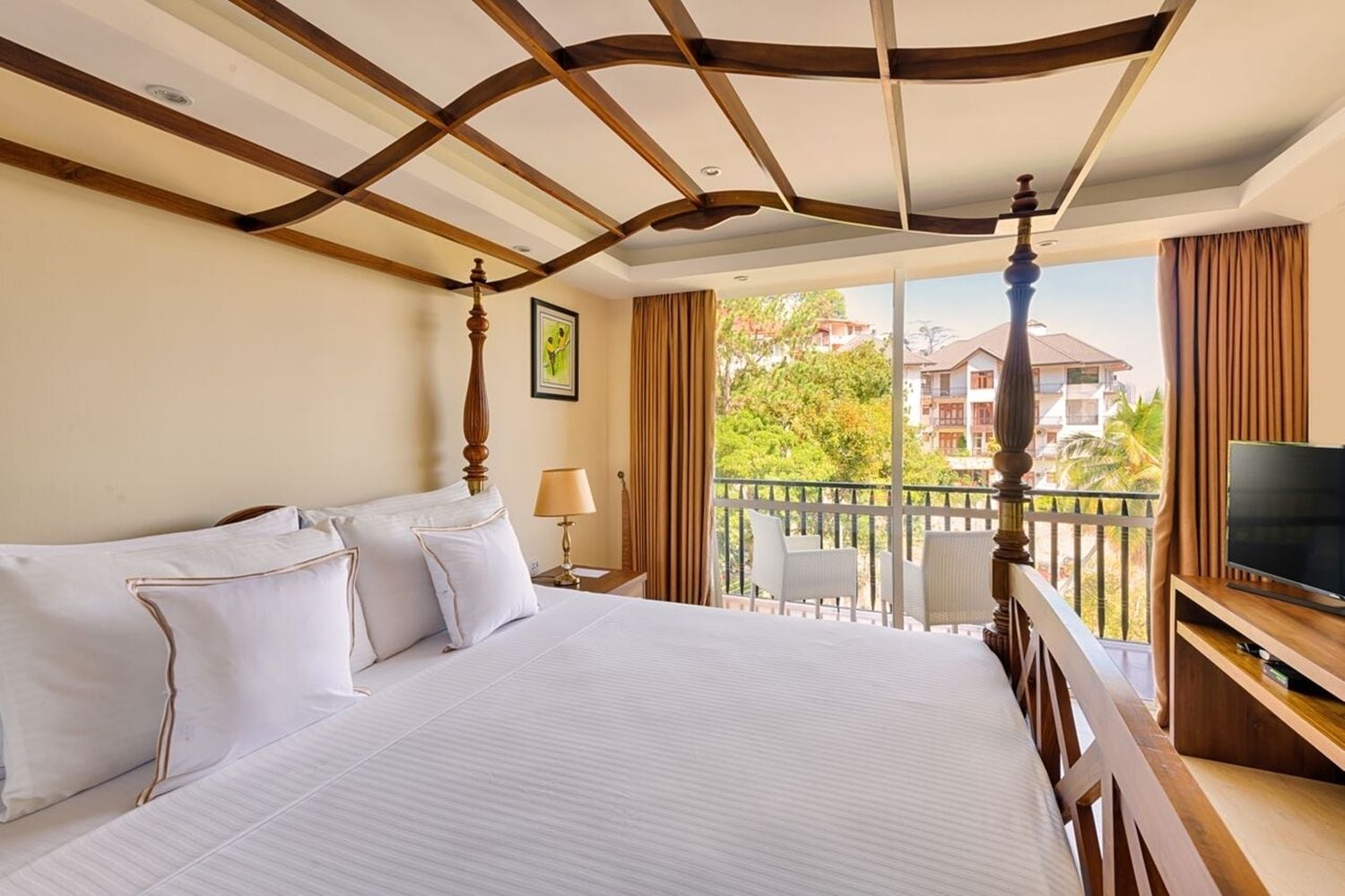 A hotel room with a large bed, a television, and a balcony with a view of buildings and trees.