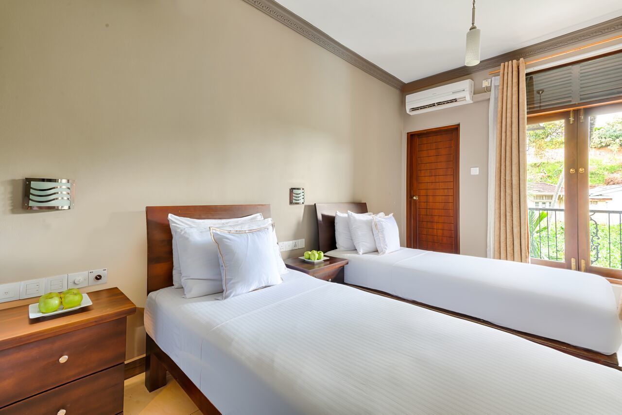 Ein Hotelzimmer mit zwei Betten, einem Nachttisch aus Holz, einem kleinen Tisch mit einem Teller Äpfeln, einer Klimaanlage und Blick auf Grün durch das Fenster.