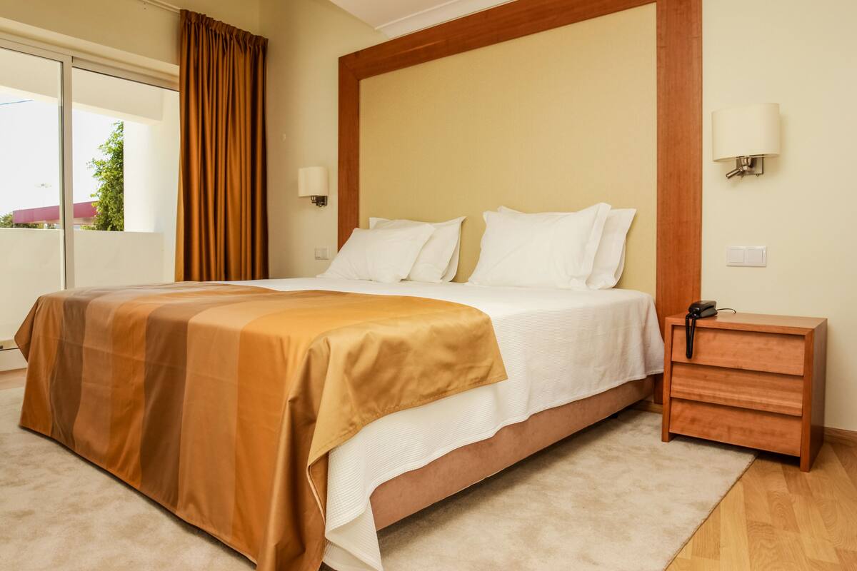 A hotel room with a large bed, a wooden nightstand, a lamp, and a view of the outdoors.