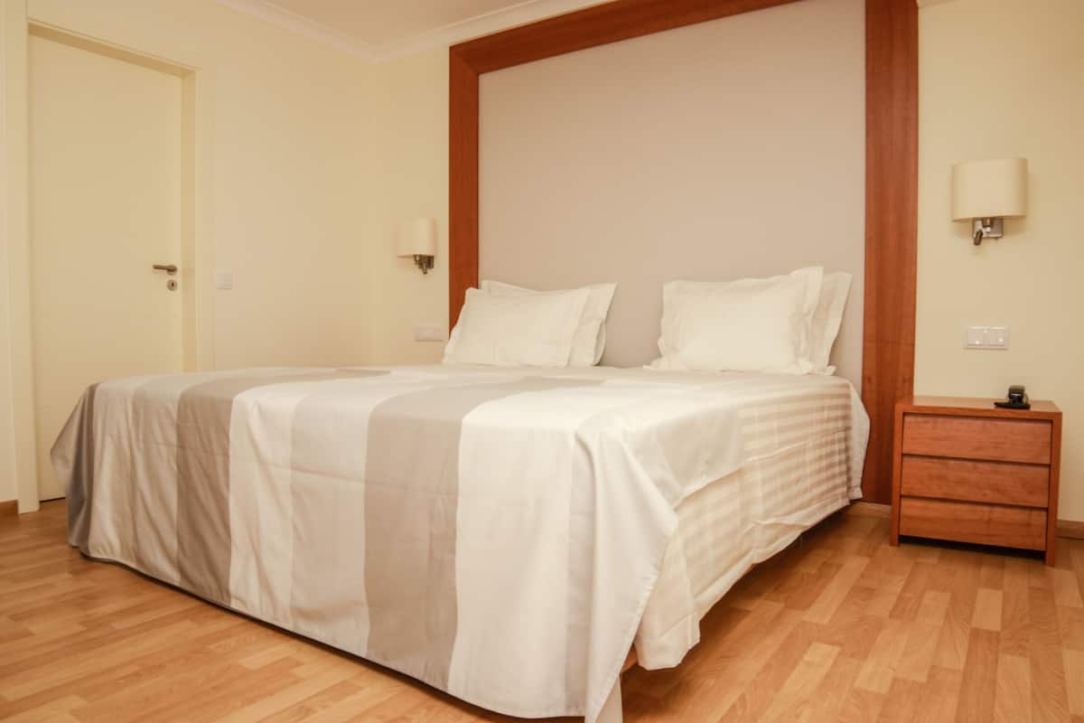 A hotel room with a large bed, wooden floor, a nightstand with a lamp, and a door.