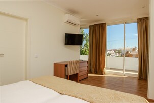 Habitación de hotel con cama, televisor en la pared, cómoda de madera y puerta corrediza de vidrio al exterior.