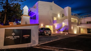 Un hotel moderno con fachada blanca, un coche estacionado frente a él y un letrero que indica que está abierto.