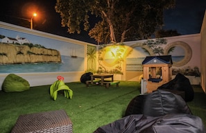 Un patio trasero con césped artificial, una casita de juegos de madera y un mural de una escena costera.