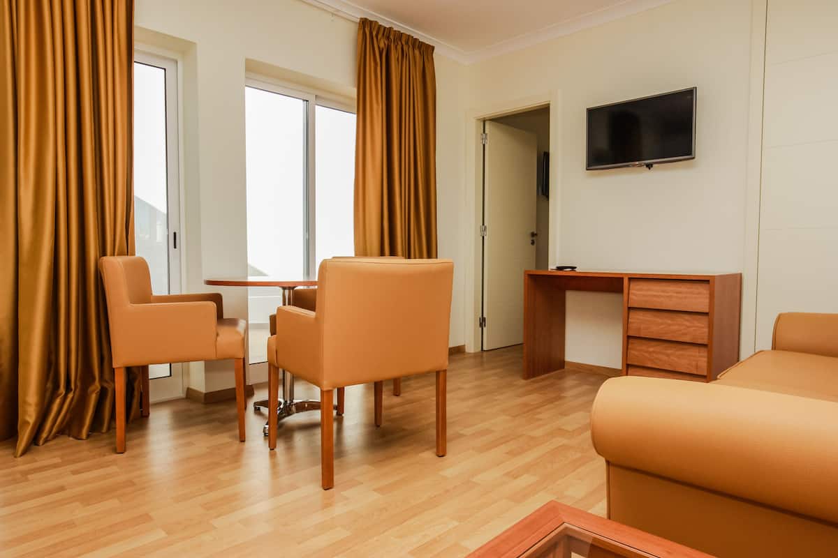 A hotel room with a wooden floor, a sofa, a desk with a chair, a television on the wall, and large windows with curtains.
