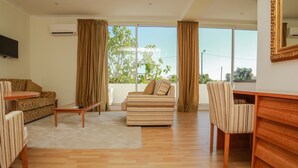 Una sala de estar con sofá, sillón y una mesita. Cuenta con un ventanal con cortinas, un televisor de pantalla plana montado en la pared y un escritorio de madera con una silla.