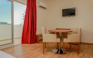 A room with a round wooden table, two beige chairs, a red curtain, an air conditioning unit, and a flat-screen TV mounted on the wall.