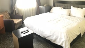 A hotel room with a large bed, a bedside table with a remote control, a chair, a nightstand, and a window with curtains. - Pentagon Luxury Suites Enugu (Enugu)
