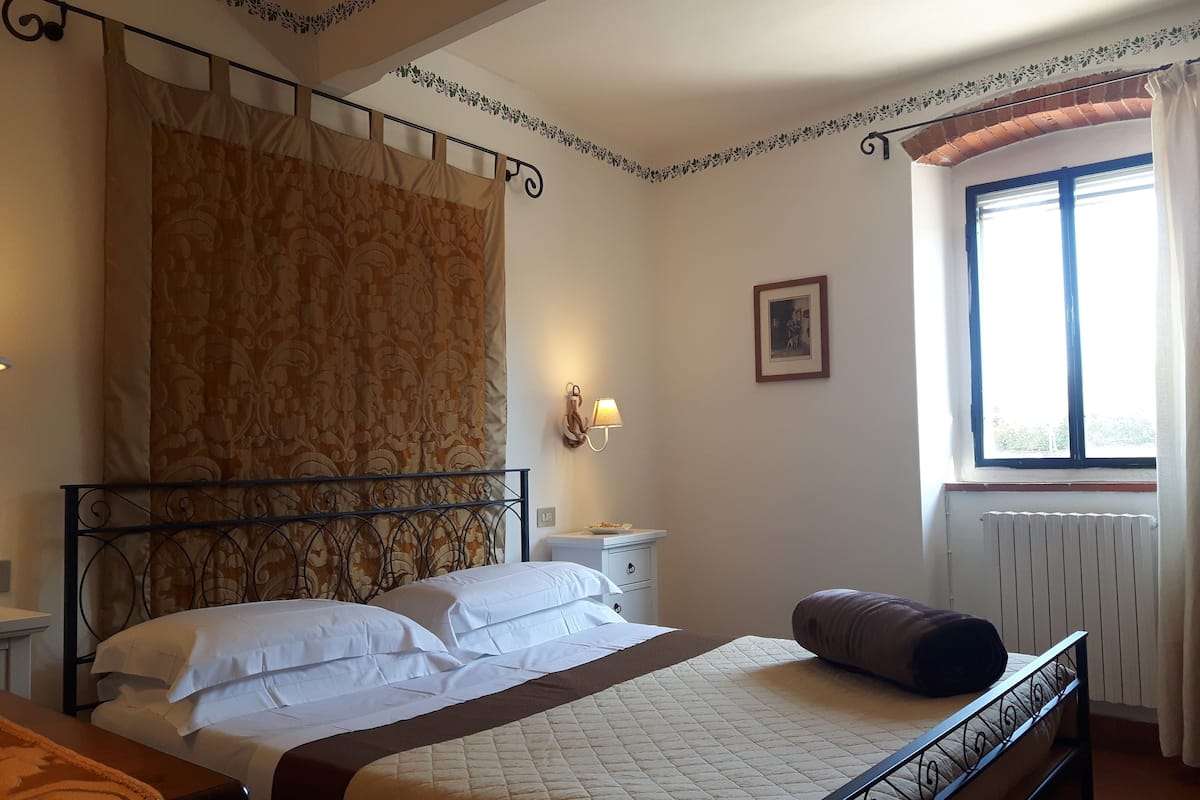 A bedroom with a bed, a nightstand, a wall-mounted lamp, a window with curtains, and a radiator.