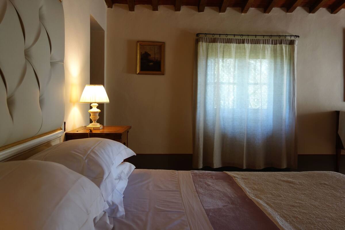 A bedroom with a bed, a nightstand with a lamp, a painting on the wall, and a window with curtains.