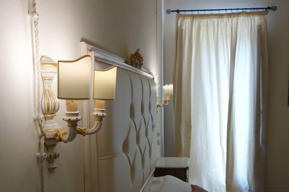 A hotel room with a classic design, featuring a large bed with white linens and two decorative pillows, a small bedside table, and wall-mounted lamps.