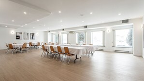 Una amplia sala de conferencias con mesas y sillas blancas, ventanas grandes y un techo alto con iluminación empotrada.