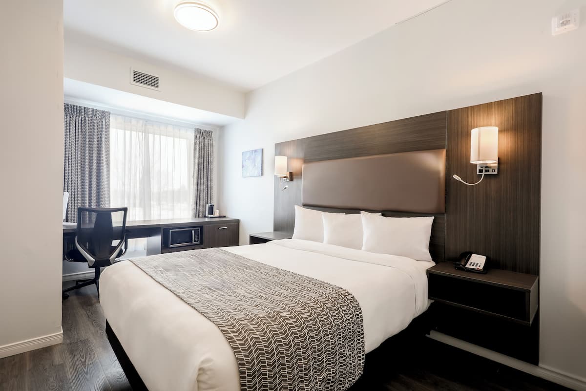 A hotel room with a large bed, a desk with a chair, a nightstand with a phone, and a window with curtains.