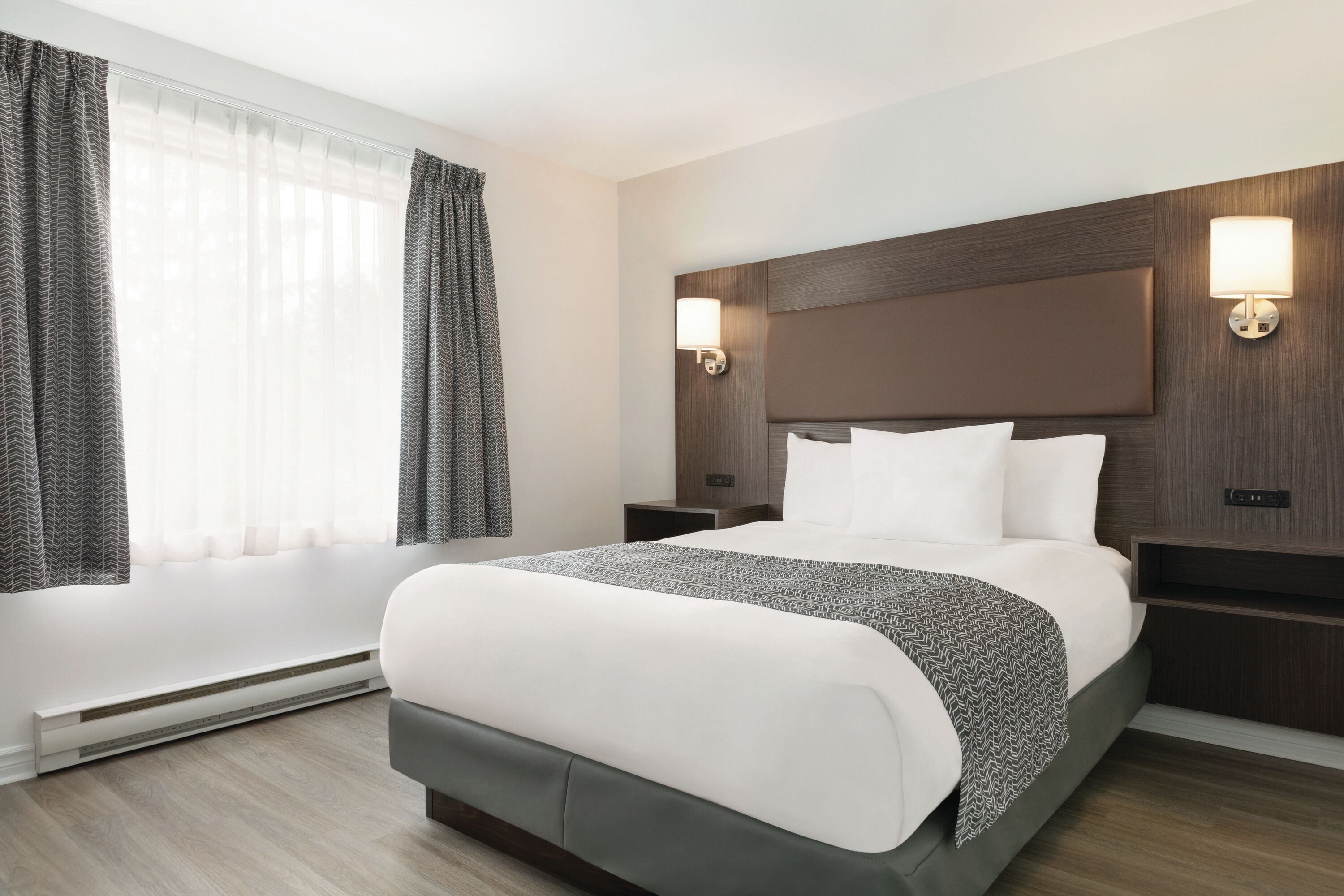 Une chambre d’hôtel avec un grand lit, deux tables de chevet, une lampe fixée au mur et une fenêtre avec des rideaux.