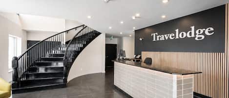 A reception area with a curved staircase, a reception desk, and a wall-mounted sign reading 'Travelodge'.