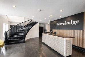 A reception area with a curved staircase, a reception desk, and a wall-mounted sign reading 'Travelodge'.