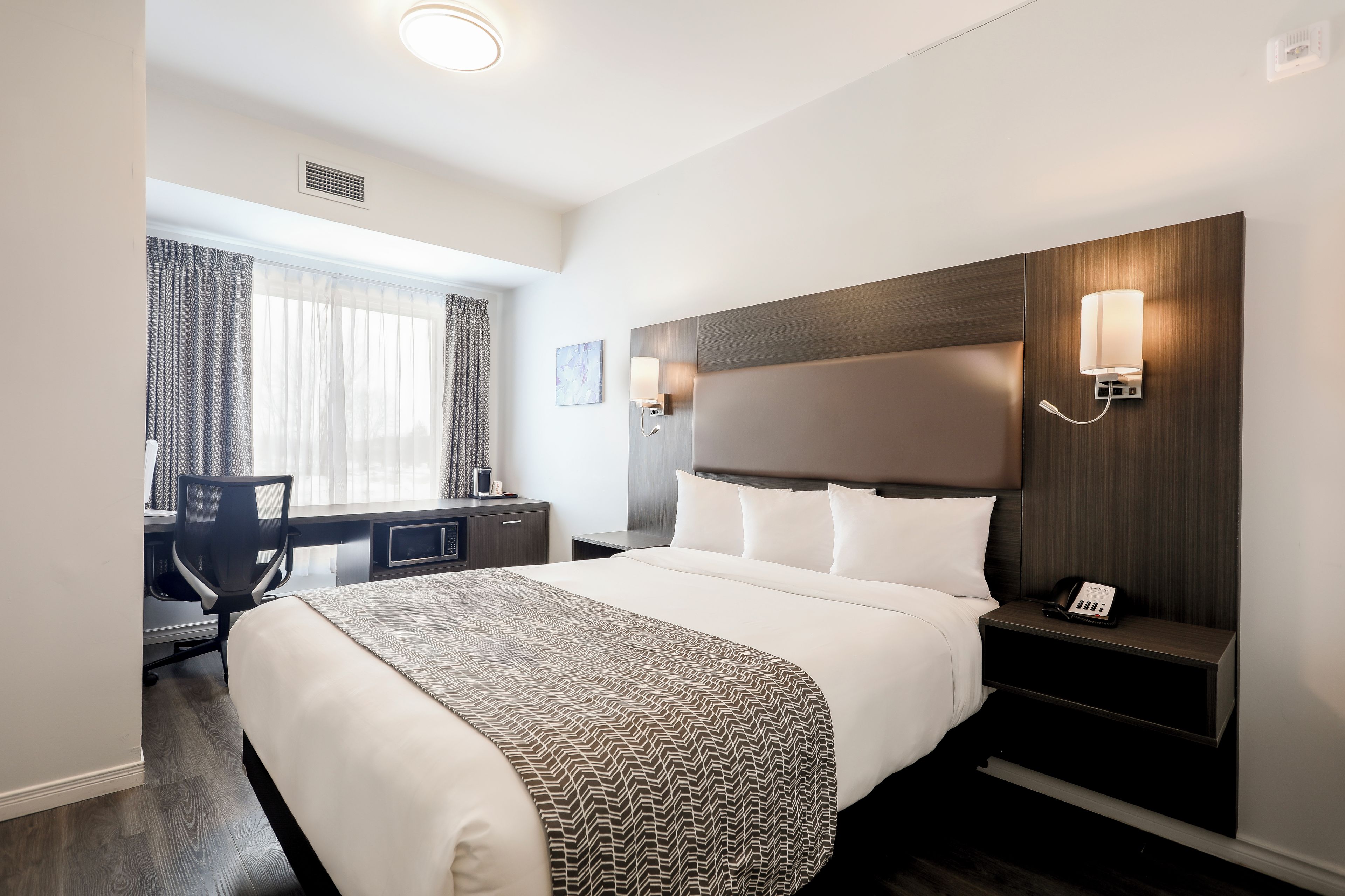 A hotel room with a large bed, a desk with a chair, a nightstand with a phone, and a window with curtains.