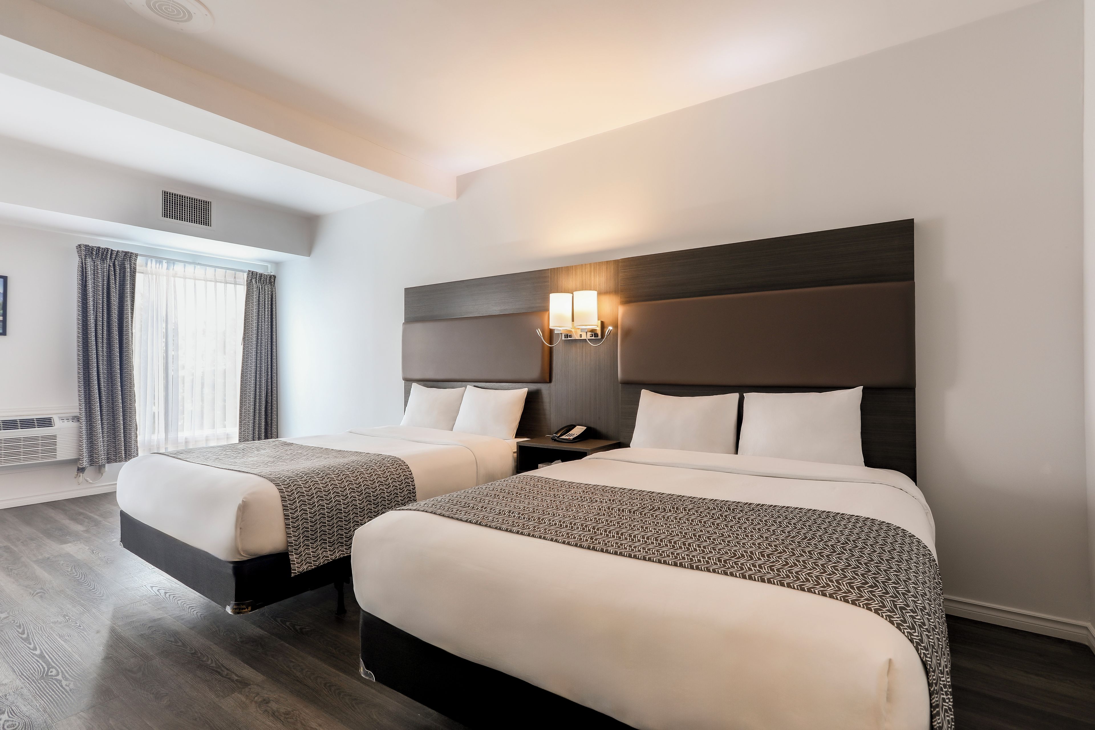 Habitación de hotel con dos camas, un ventanal con cortinas, una lámpara fijada a la pared y suelo de madera.