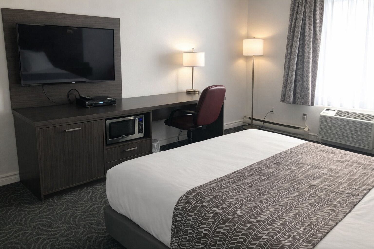 A hotel room with a bed, a desk with a TV, a microwave, a chair, and two lamps.