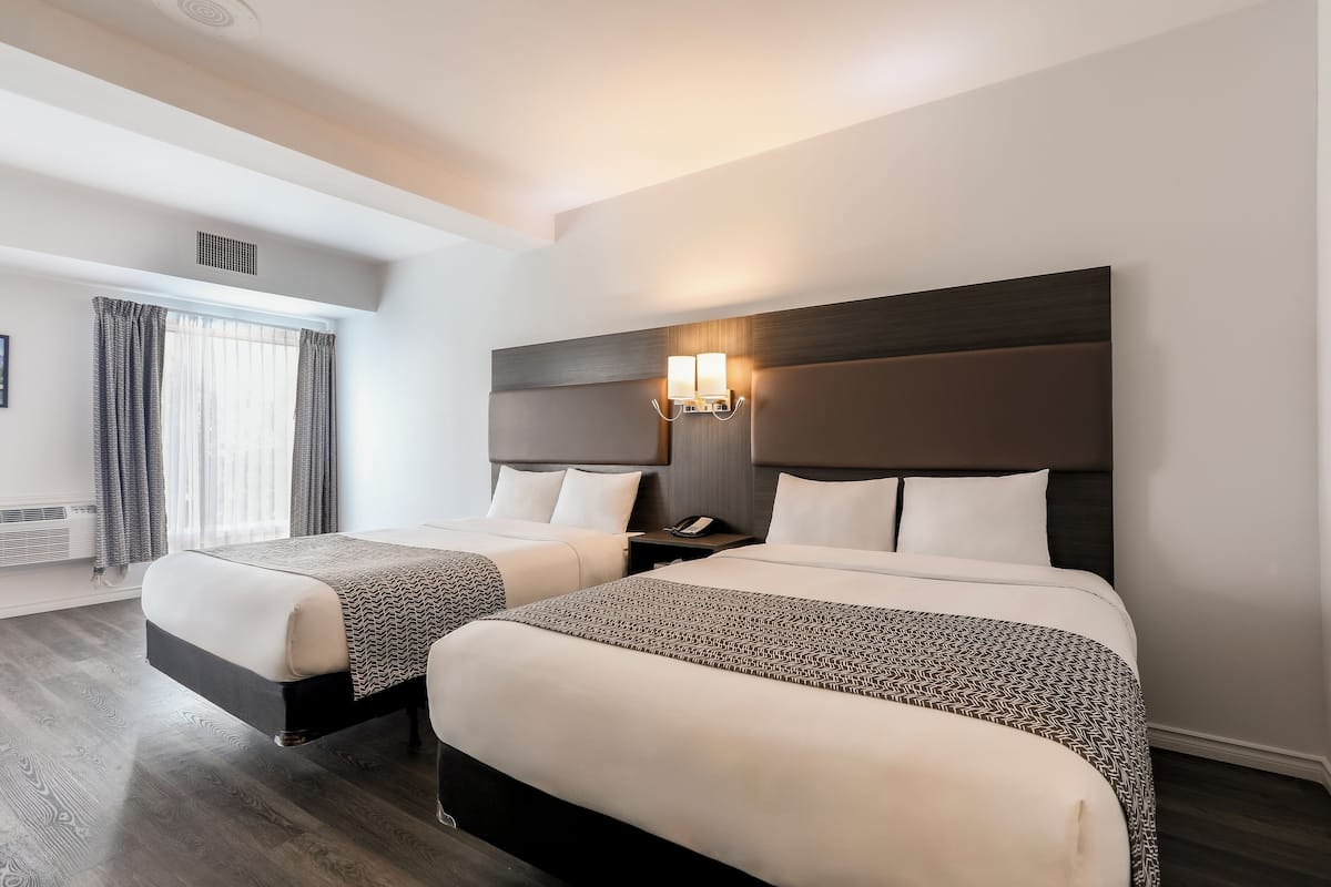 A hotel room with two beds, a large window with curtains, a wall-mounted light fixture, and a wooden floor.