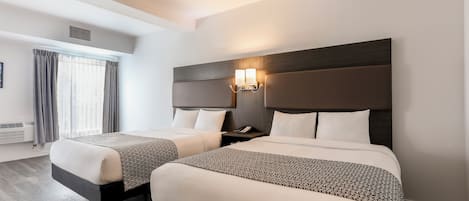 A hotel room with two beds, a large window with curtains, a wall-mounted light fixture, and a wooden floor.