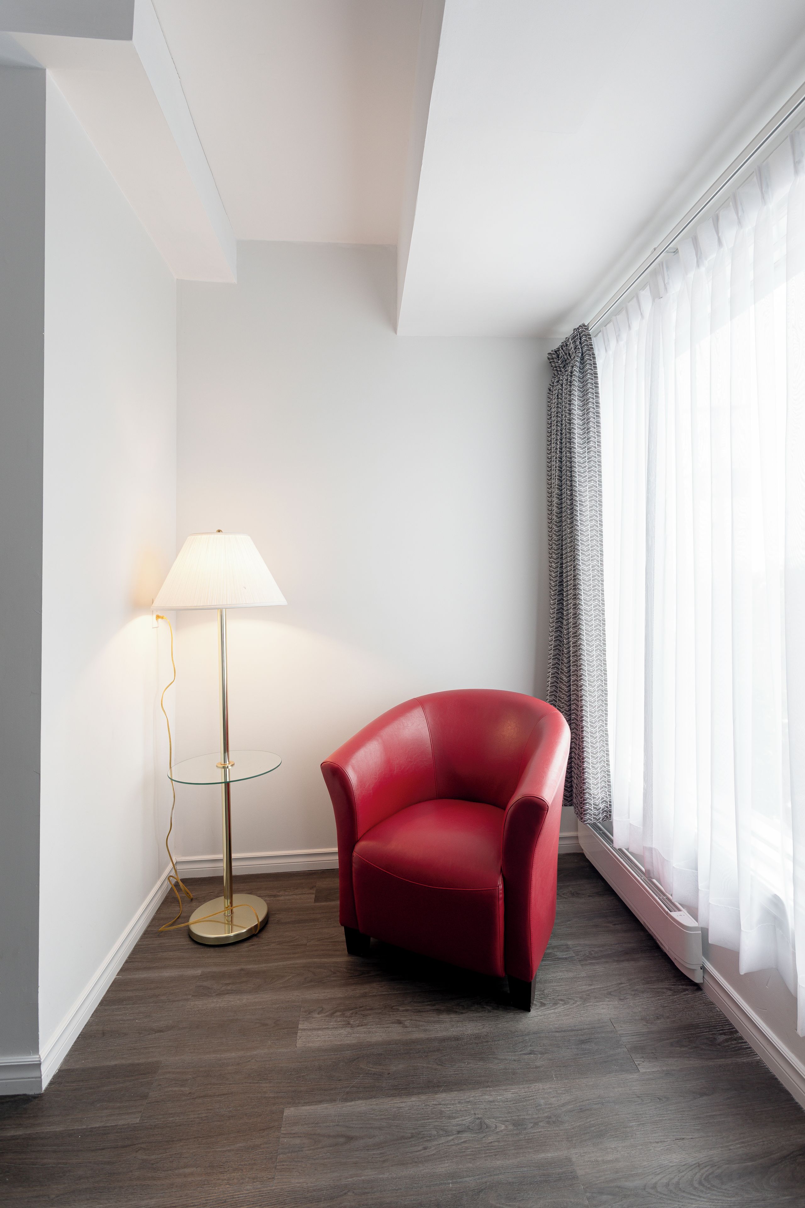 A corner room with a red chair, a floor lamp, and a large window with sheer curtains.