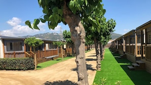 A row of wooden cabins with a dirt path and green lawn, surrounded by trees and hills in the background. - Camping El Camell (Pineda de Mar)