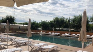 A poolside area with white lounge chairs and beige umbrellas, surrounded by trees and a building in the background. - Camping El Camell (Pineda de Mar)